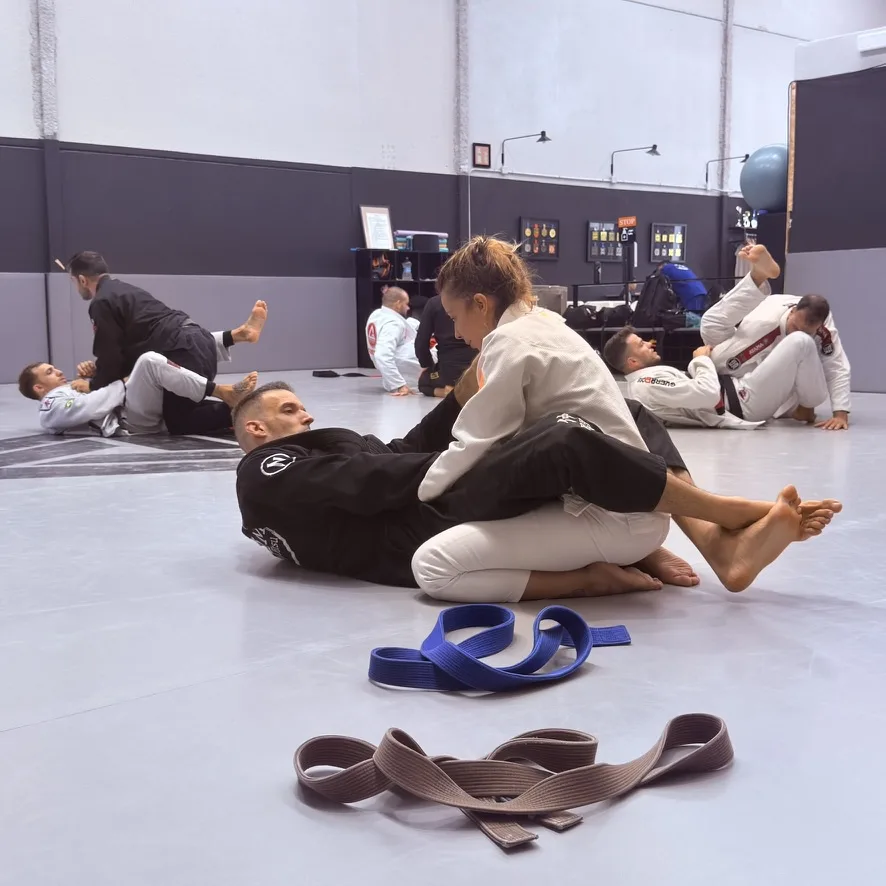 Una alumna de Elite BJJ controla a su compañero en el suelo durante la clase, con cinturones azul y marrón en primer plano