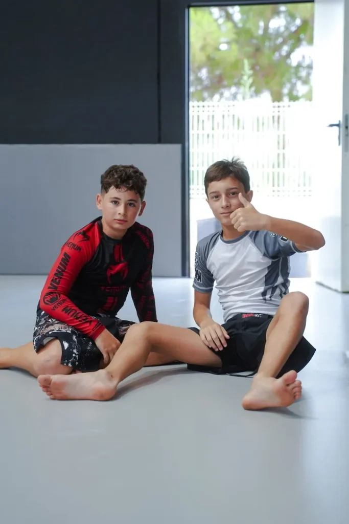 Dos chicos de Elite BJJ sonrientes en el tatami después de entrenar No-Gi