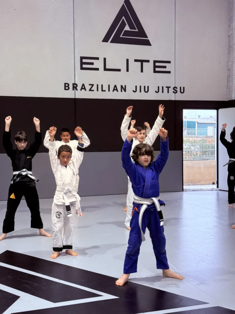 Grupo de niños con kimono calentando con los brazos arriba en el tatami de Elite BJJ